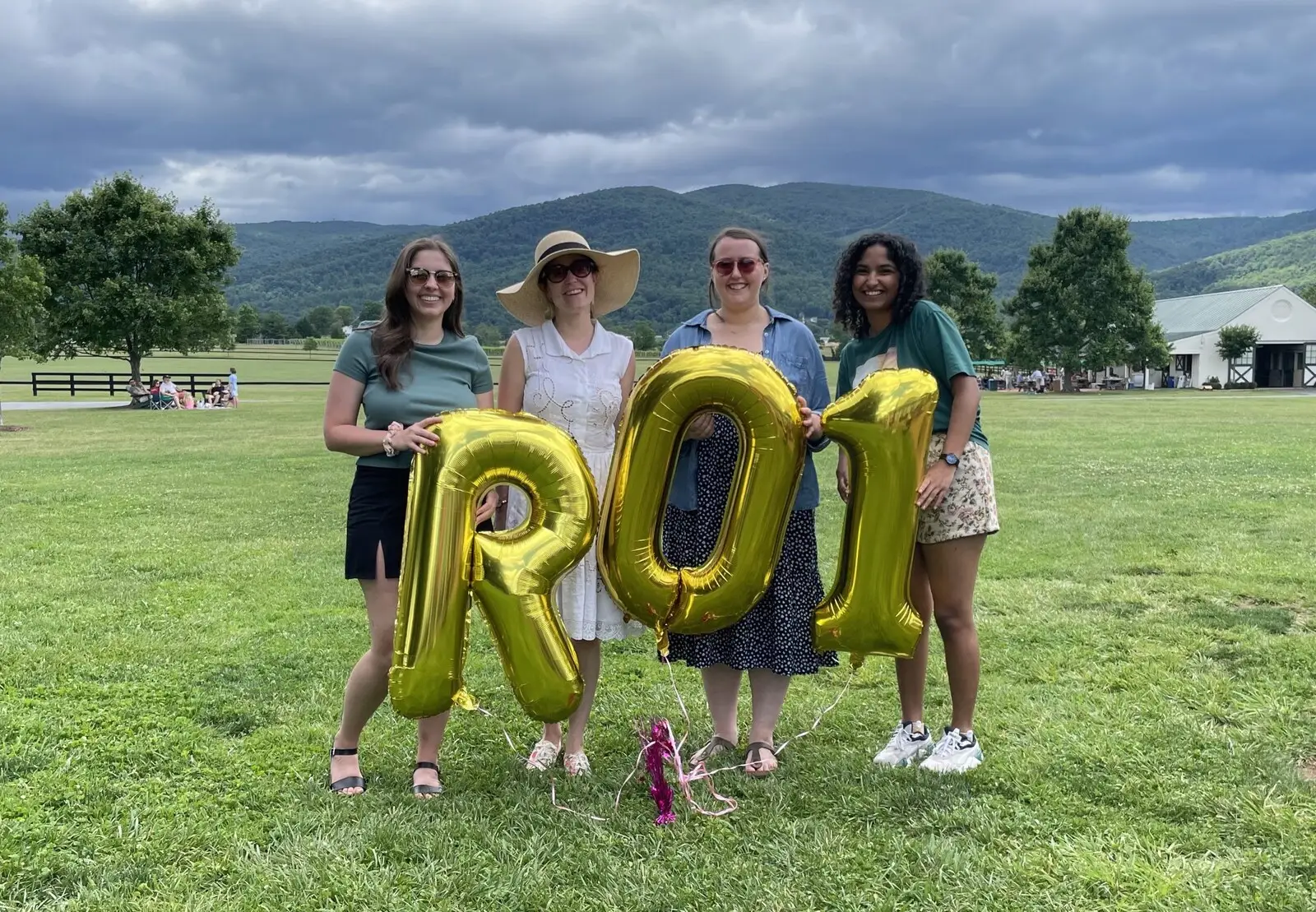Four lab members on a lawn holding gold balloons spelling R01, with hills behind them
