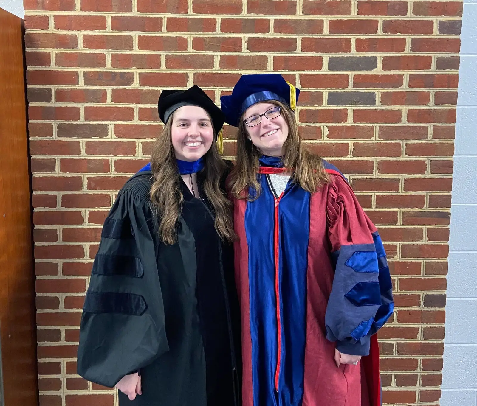 Two people in doctoral gowns and caps outside a brick building